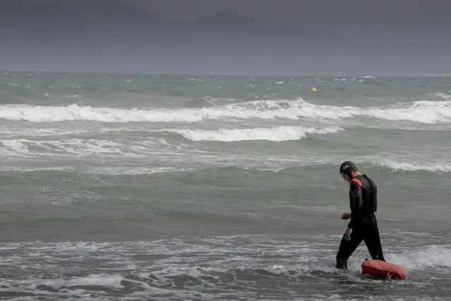 Ein Surfer mit rotem Board geht in die Brandung, im Hintergrund liegen Berge.